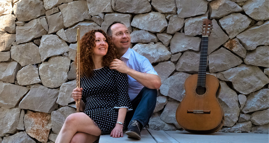Cavatina Duo seated against a stone wall, the flutist holding her flute beside a classical guitar