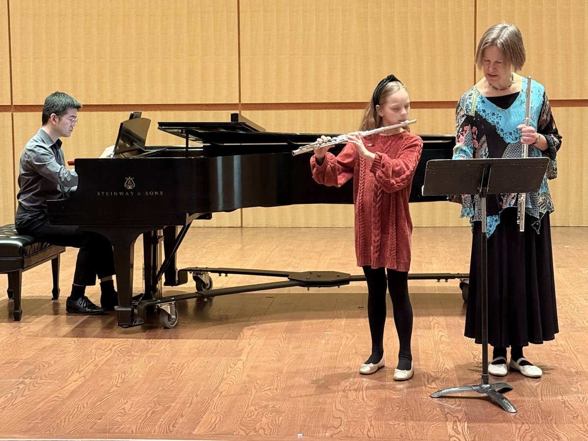 A young flutist performing on stage with a piano accompanist and her teacher turning pages at the music stand
