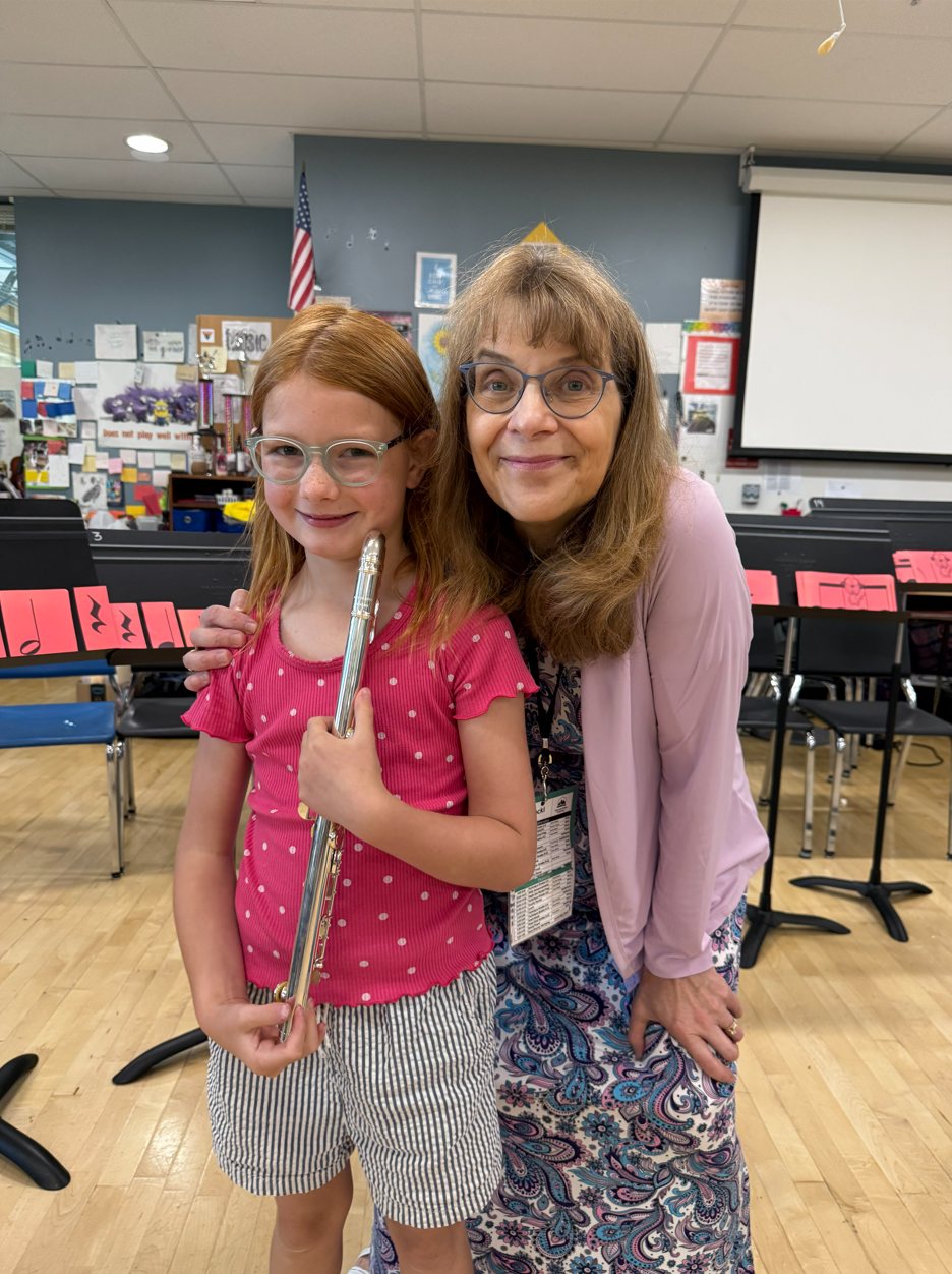 A young flute student holding her flute stands beside her smiling teacher in a school band room